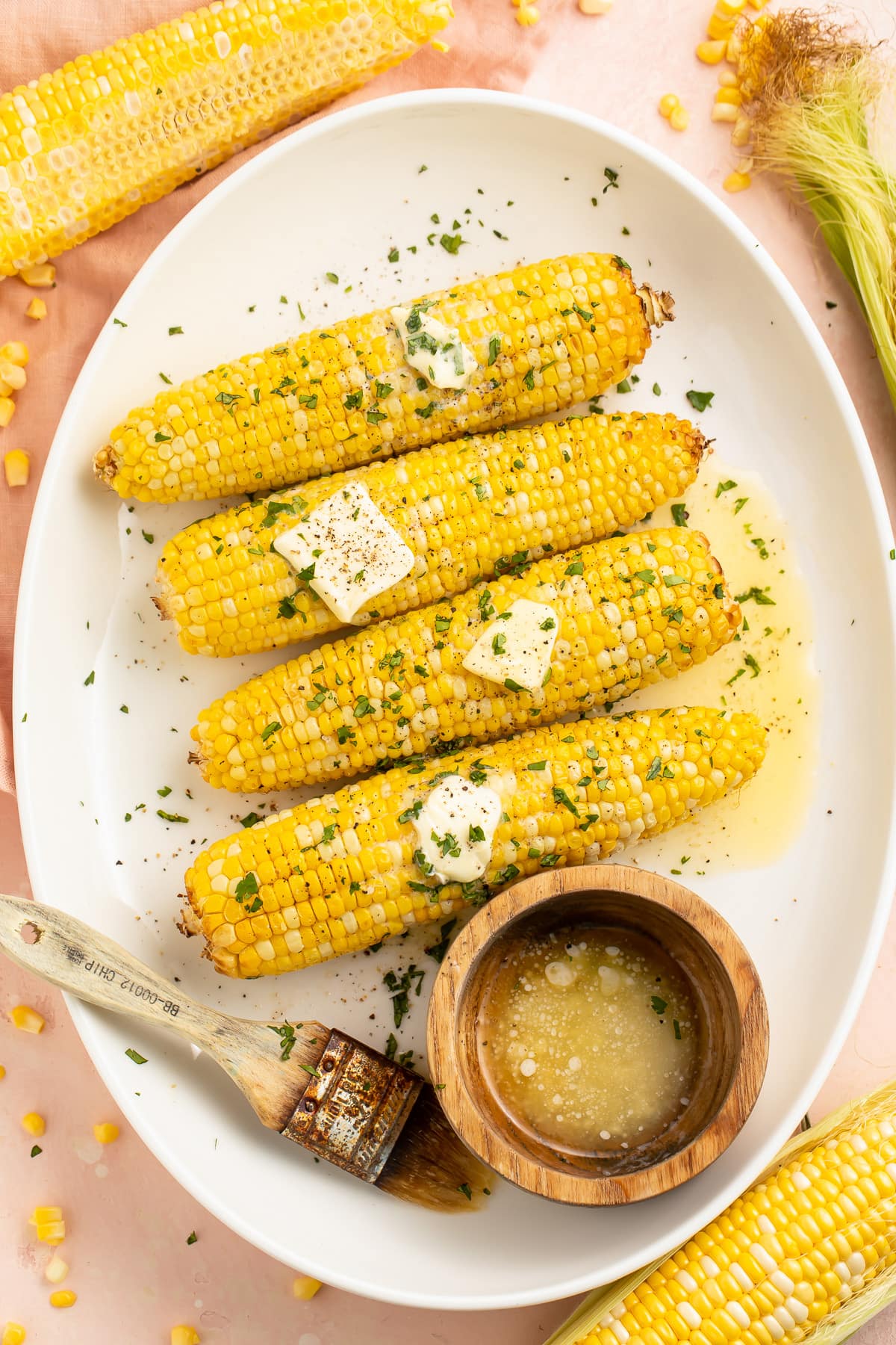 4 ears of air fryer corn on the cob arranged on a white oval platter next to a ramekin of melted butter.