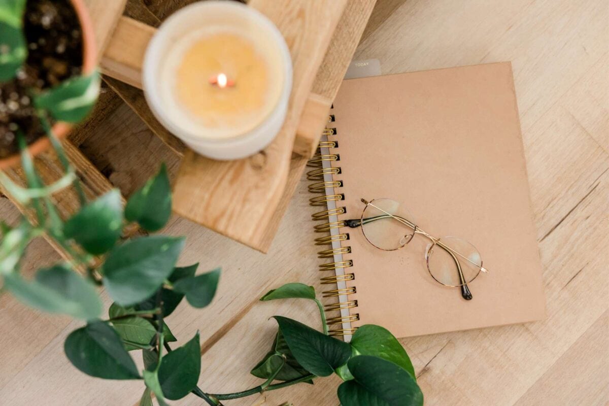 A pair of round glasses resting atop a spiral notebook next to a single wick candle and green leaves.