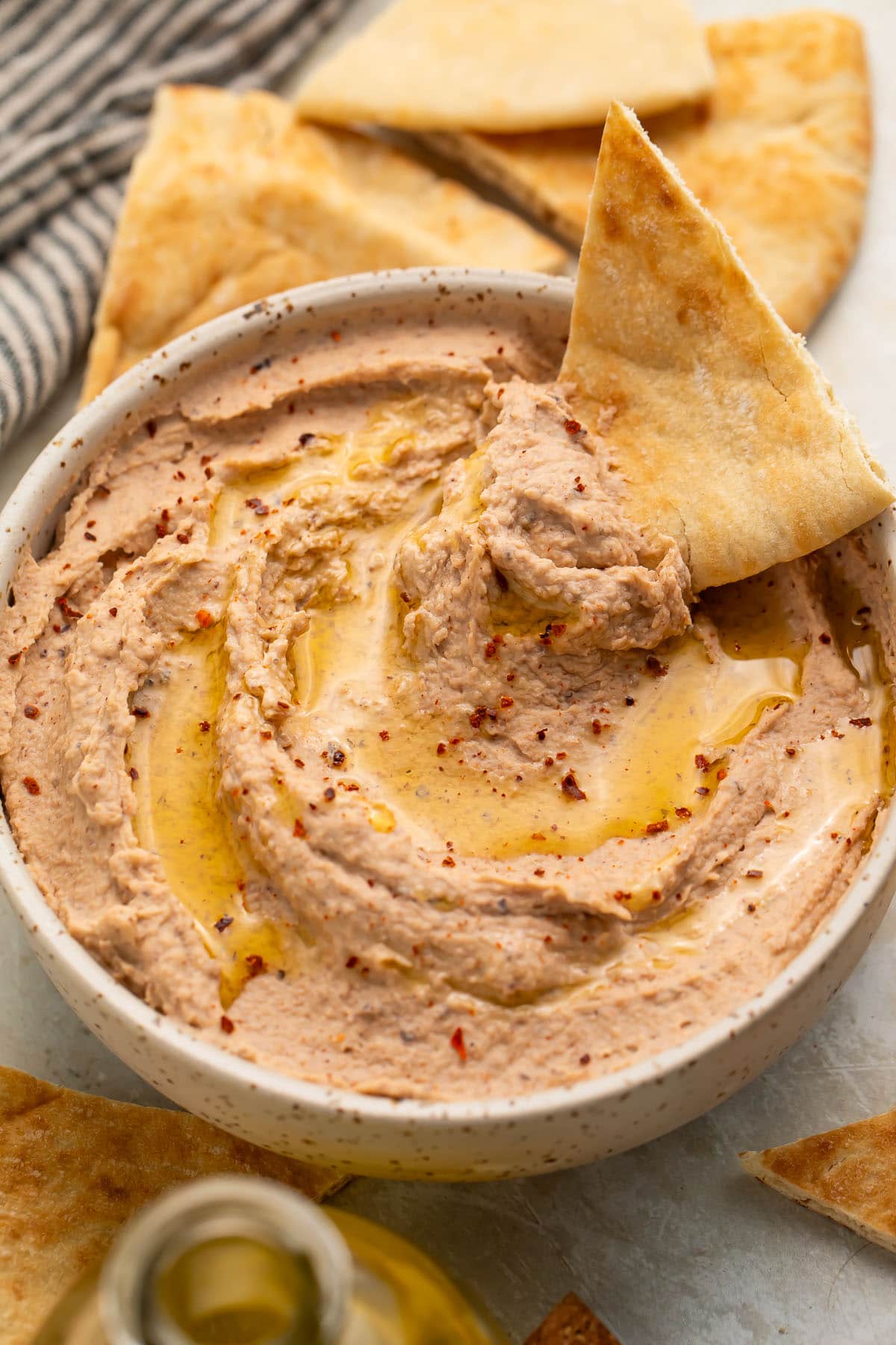 A triangular pita chip resting in a bowl of black eyed pea hummus, with a scoop of hummus on the chip.